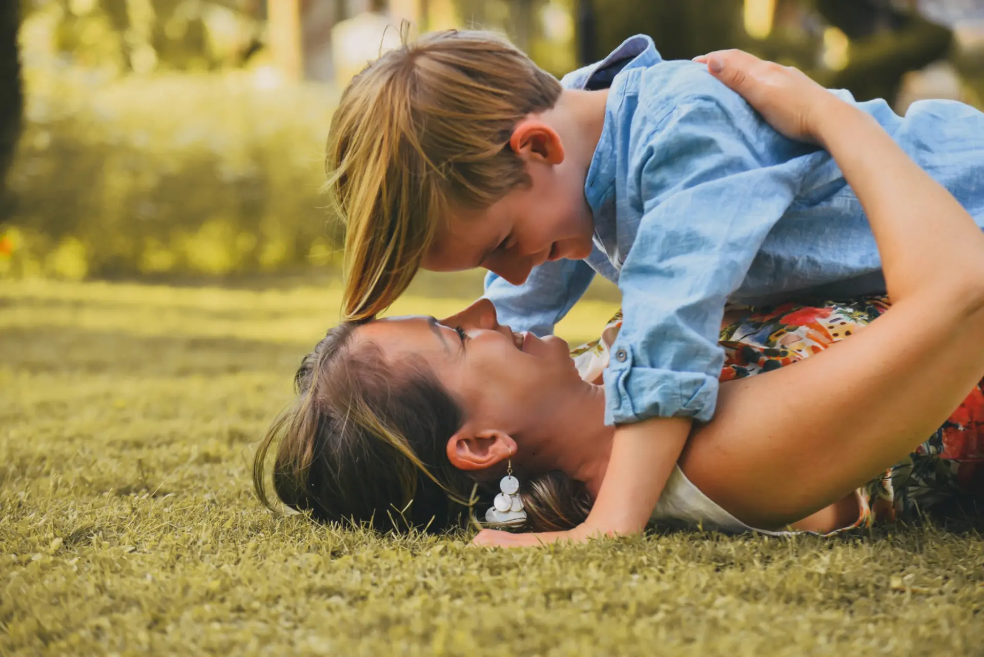 Mutter liegt im Gras, ihr Kind beugt sich lachend zu ihr herunter – enger Blickkontakt